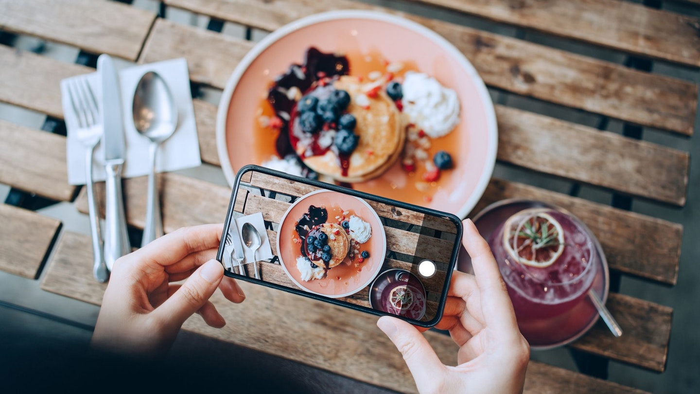 Overhead view of young Asian woman taking photos of delicious freshly served pancakes with blueberries and pink lemonade with smartphone before eating it in cafe. Eating out lifestyle. Camera eats first culture