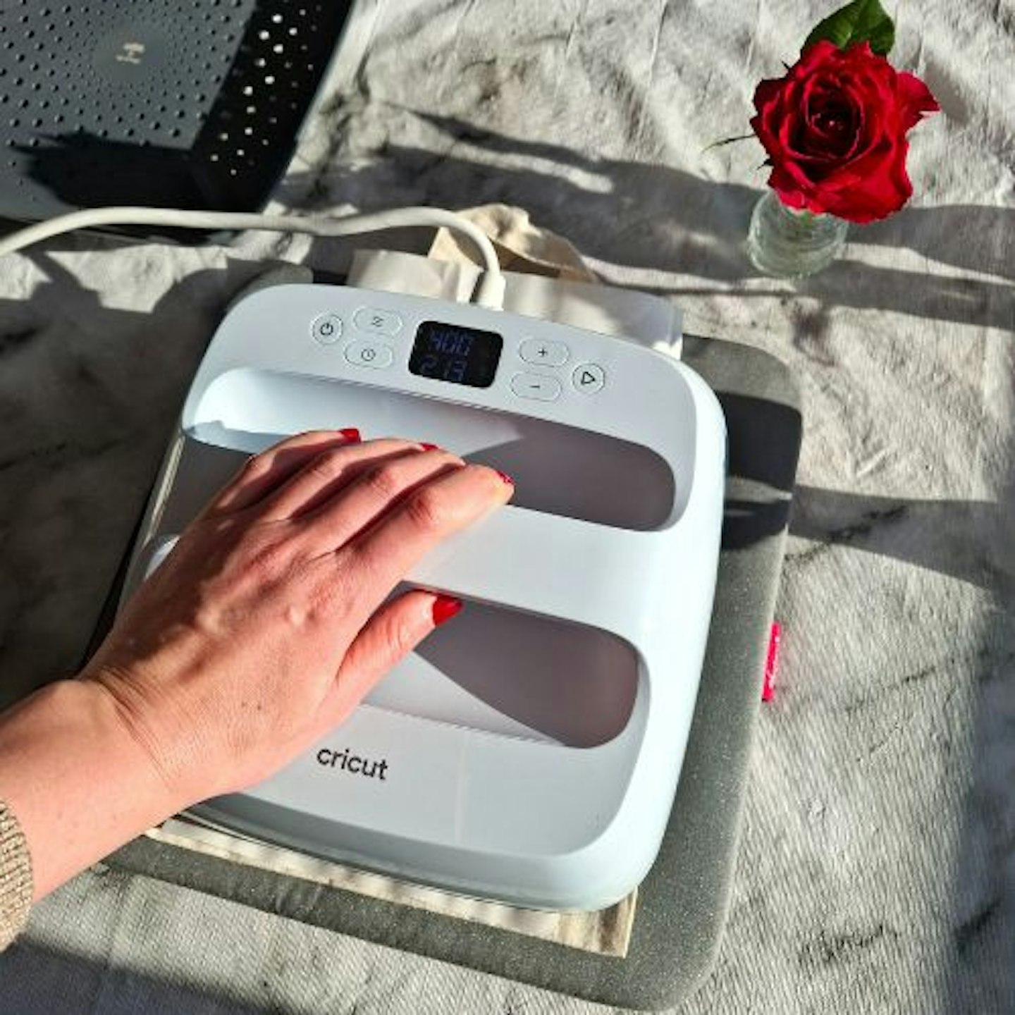 Tester's hand, with red finger nails, on the Cricut EasyPress 3. Red rose on a tabletop in background