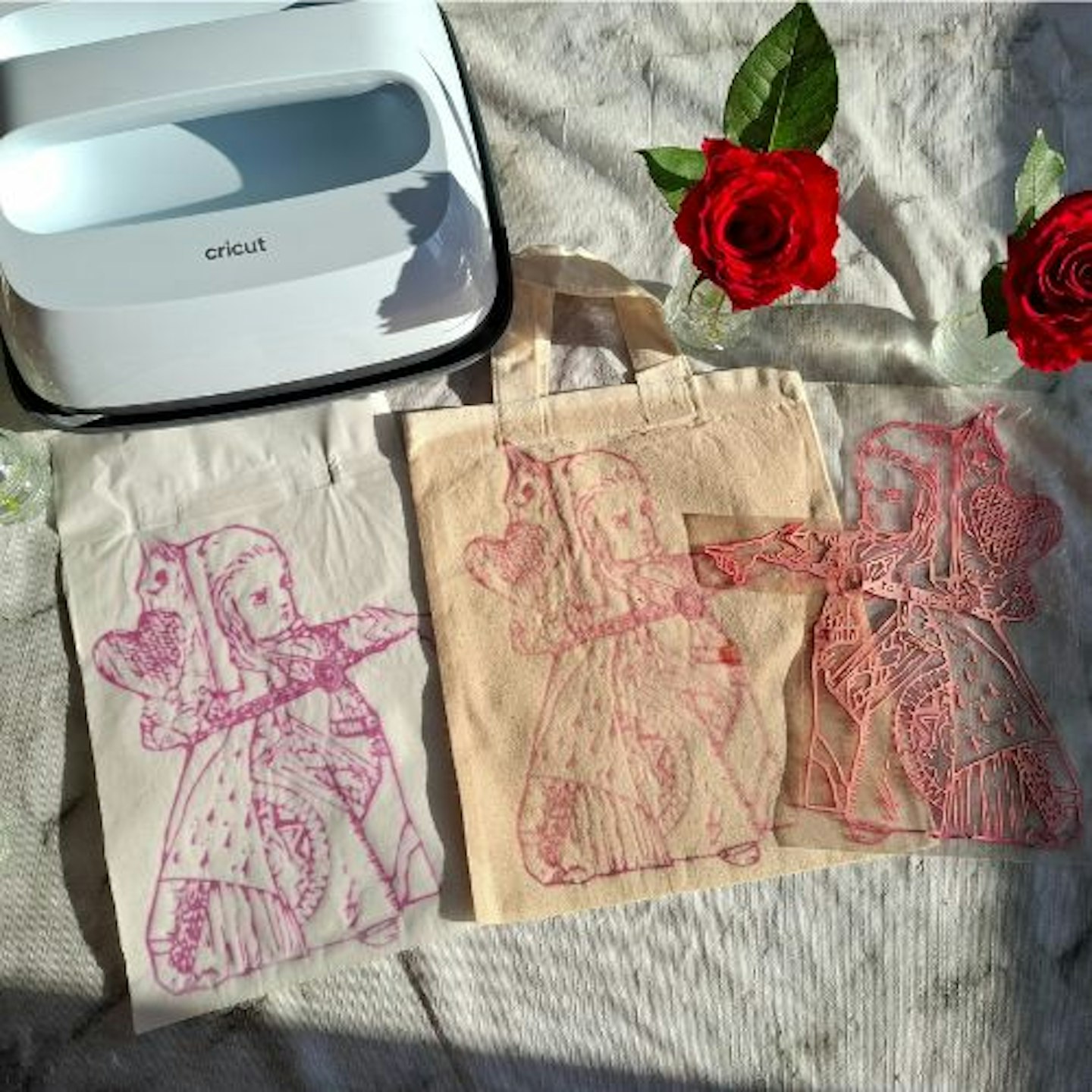 Tabletop with Cricut EasyPress 3, Natalie's Alice in Wonderland design transferred to a pale fabric and a tote bag, alongside the transfer sheet. Red roses in the background