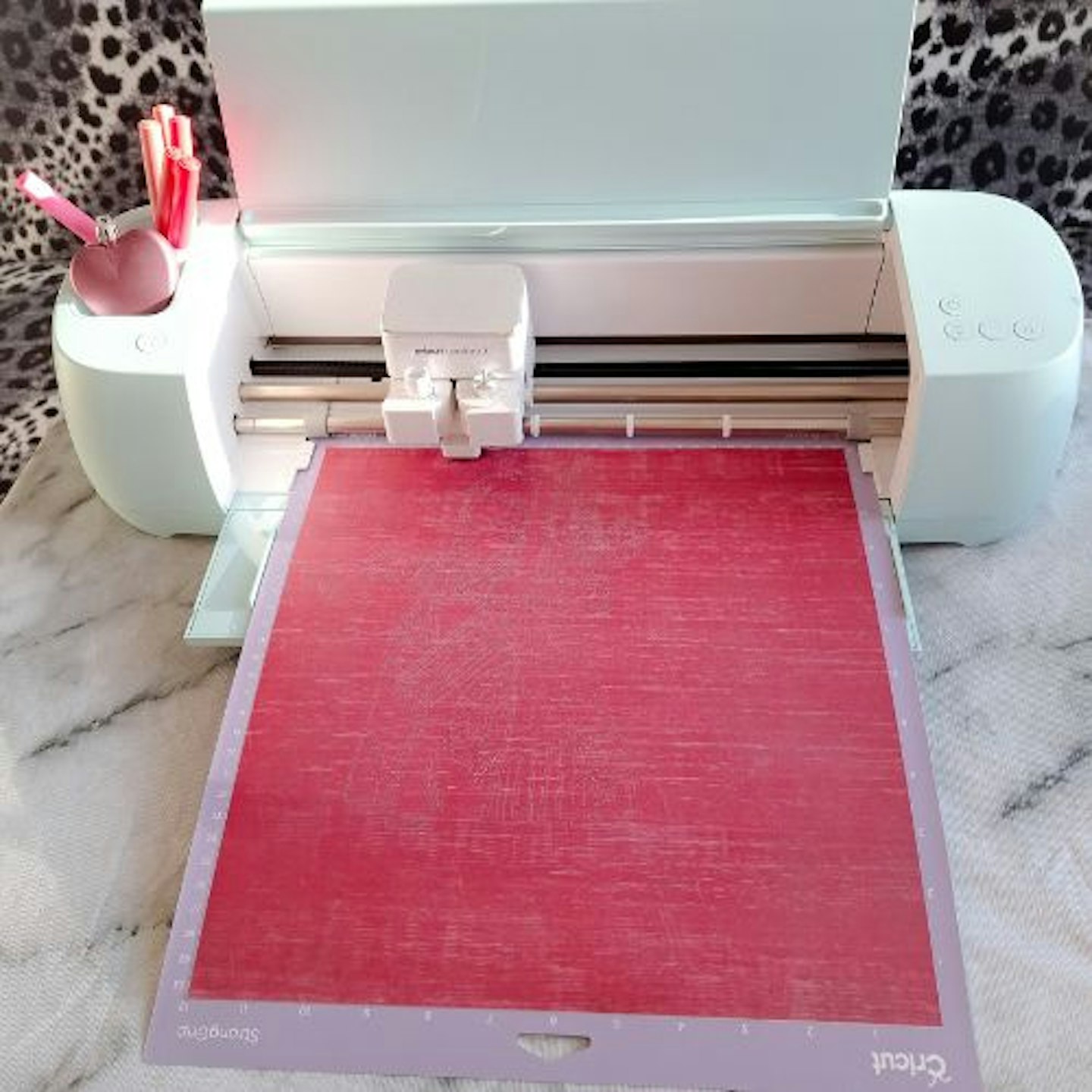 top view photo of a Cricut Explore 3 loaded with a cutting mat and pink pens in its holder on a marble design table cloth plus a monochrome leopard print background