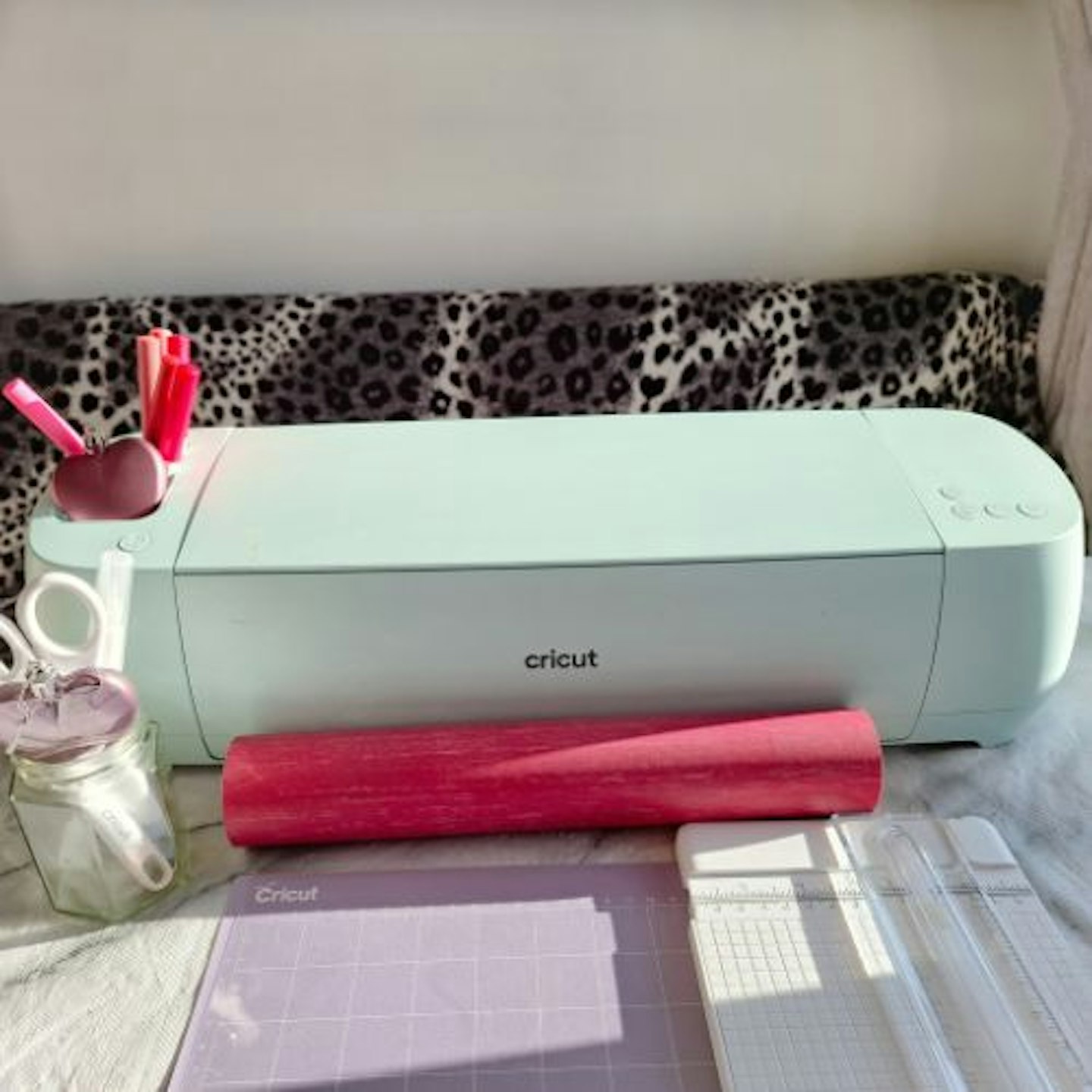 Side view of the Cricut Explore 3 in strong sunshine, surrounded by Cricut accessories and pink pens