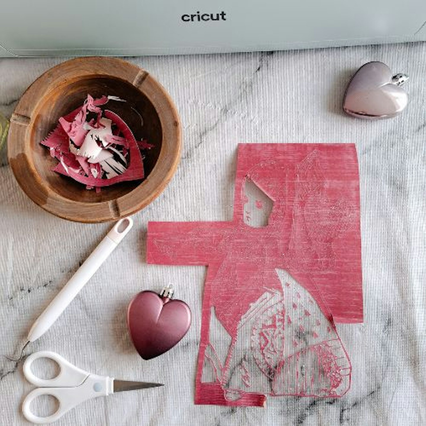 Aerial view of a half weeded design cut using the Cricut Explore 3, surrounded by weeding tools, heart baubles and a Cricut machine