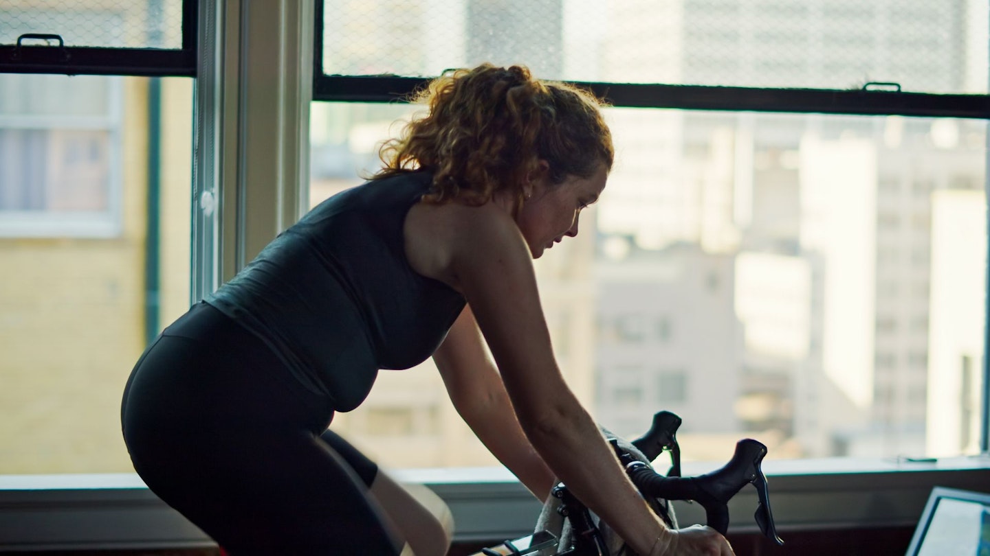 A caucasian woman in her 30s works out on a bike trainer in an urban apartment.