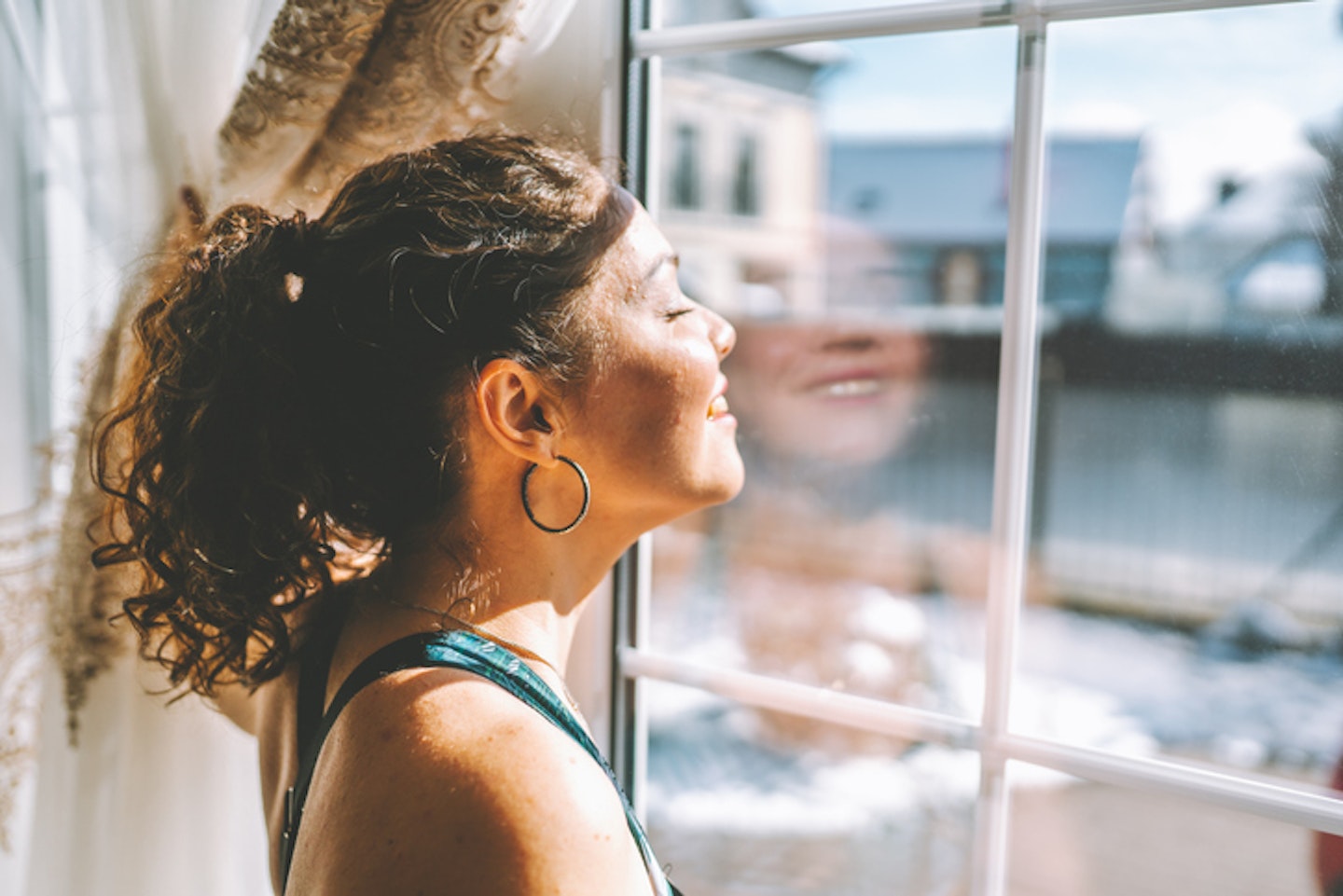 Middle age woman wearing sportswear is standing by the large window and exposing her face to the sun