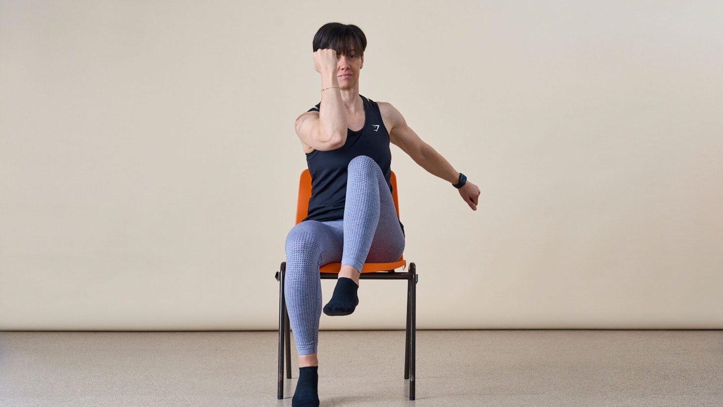 Woman doing a seated corss crunh. She is facing the camera sitting in a chair and bringing her opposite knee to elbow