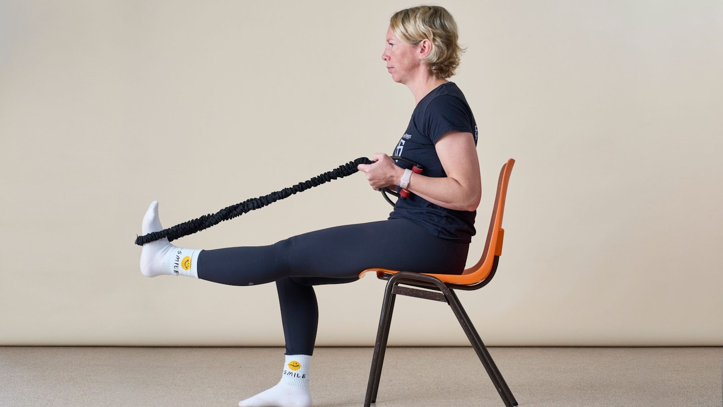 Woman sitting in a chair, side on to the camera. She is stretching her hamstring using a resistance band looped around her foot