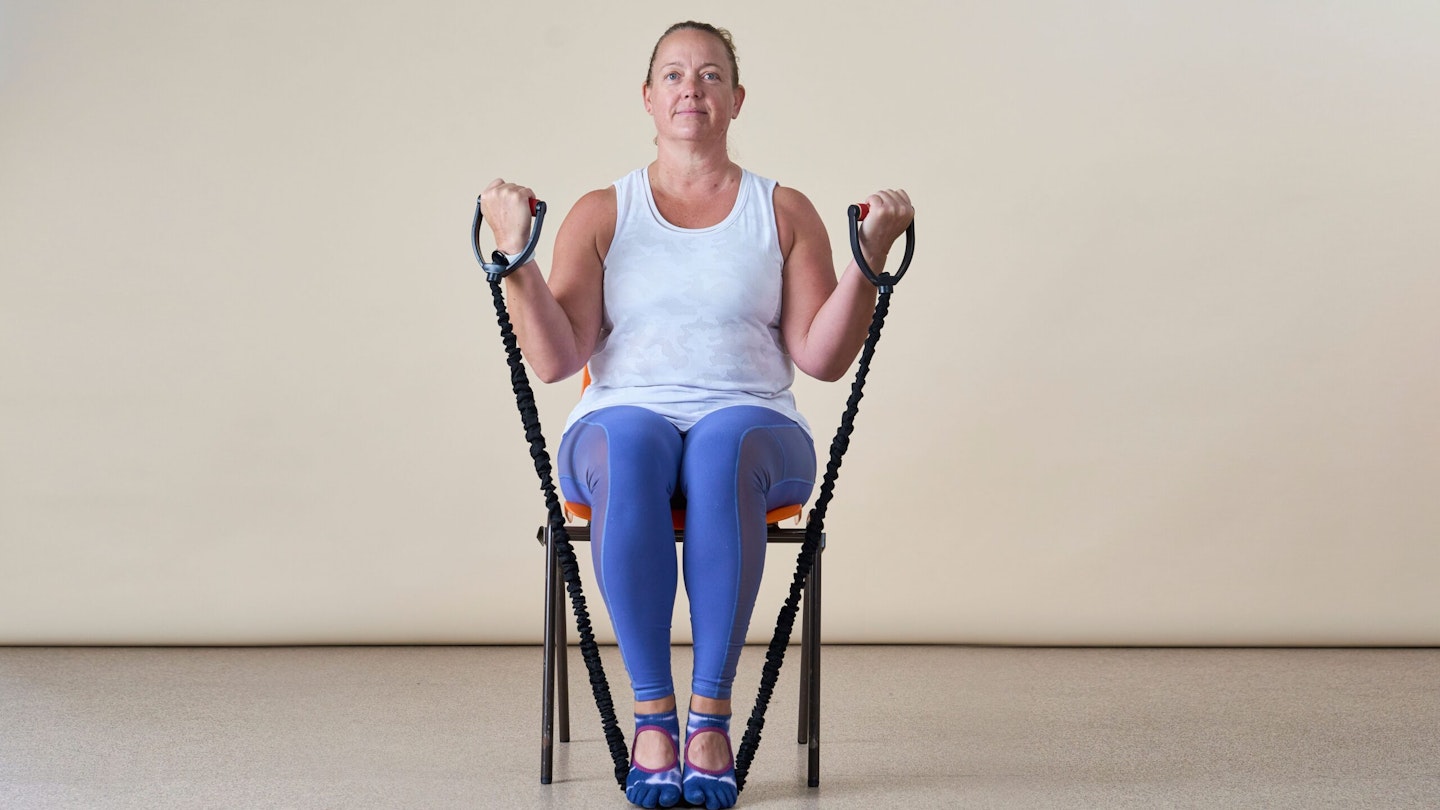 Woman facing the camera, sitting in a chair, doing a resistance band bicep curl