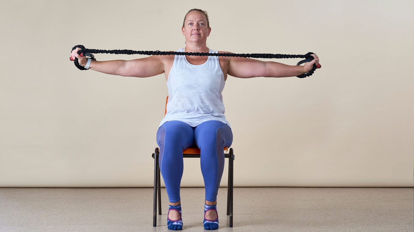 Woman facing the camera, sitting in a chair, pulling a resistance band wide