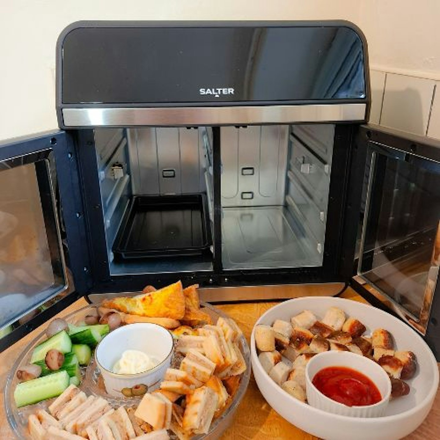 photo of the Salter French-Door Style XL Air Fryer Oven with doors wide open to show the internal cooking space, divided, with one shelf in. In the foreground are party foods that have been air fried