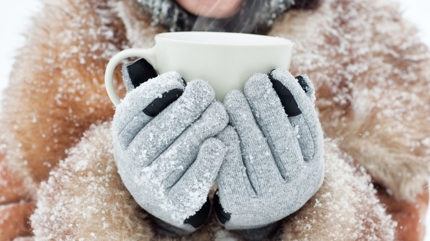 Person wearing heated gloves and holding a mug, whilst covered in snow