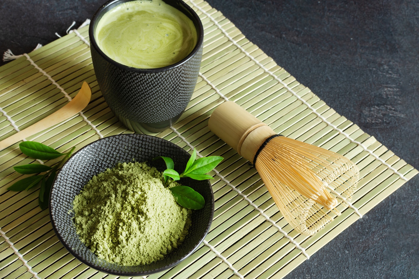 Organic green matcha tea and tea accessories on japanese mat on black background.