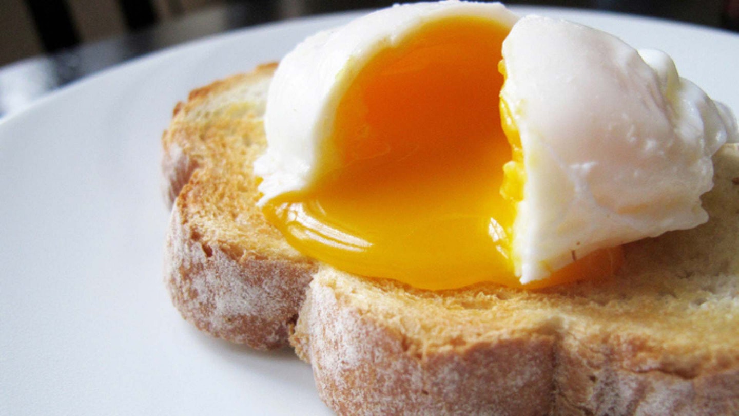 A single poached egg on artisan hand sliced toasted bread. The egg is split to reveal a runny yolk. White dinner plate, non-descript dark wood table and chair just about visible in the background.