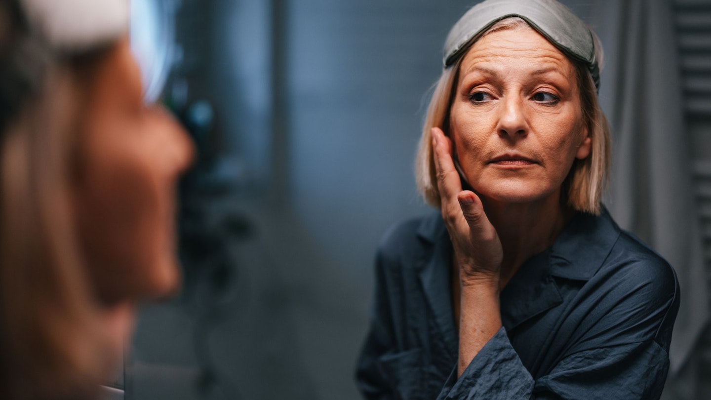 Portrait of a beautiful older woman in her pajamas and with a sleeping mask on her forehead, looking at her reflection in the mirror and checking her face and wrinkles.