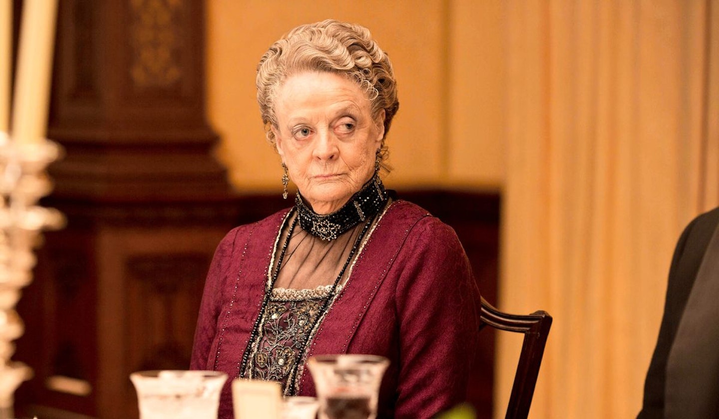 Violet Dowager (Maggie Smith) is sat on a chair at a table which has glasses on, with a contempt look on her face.