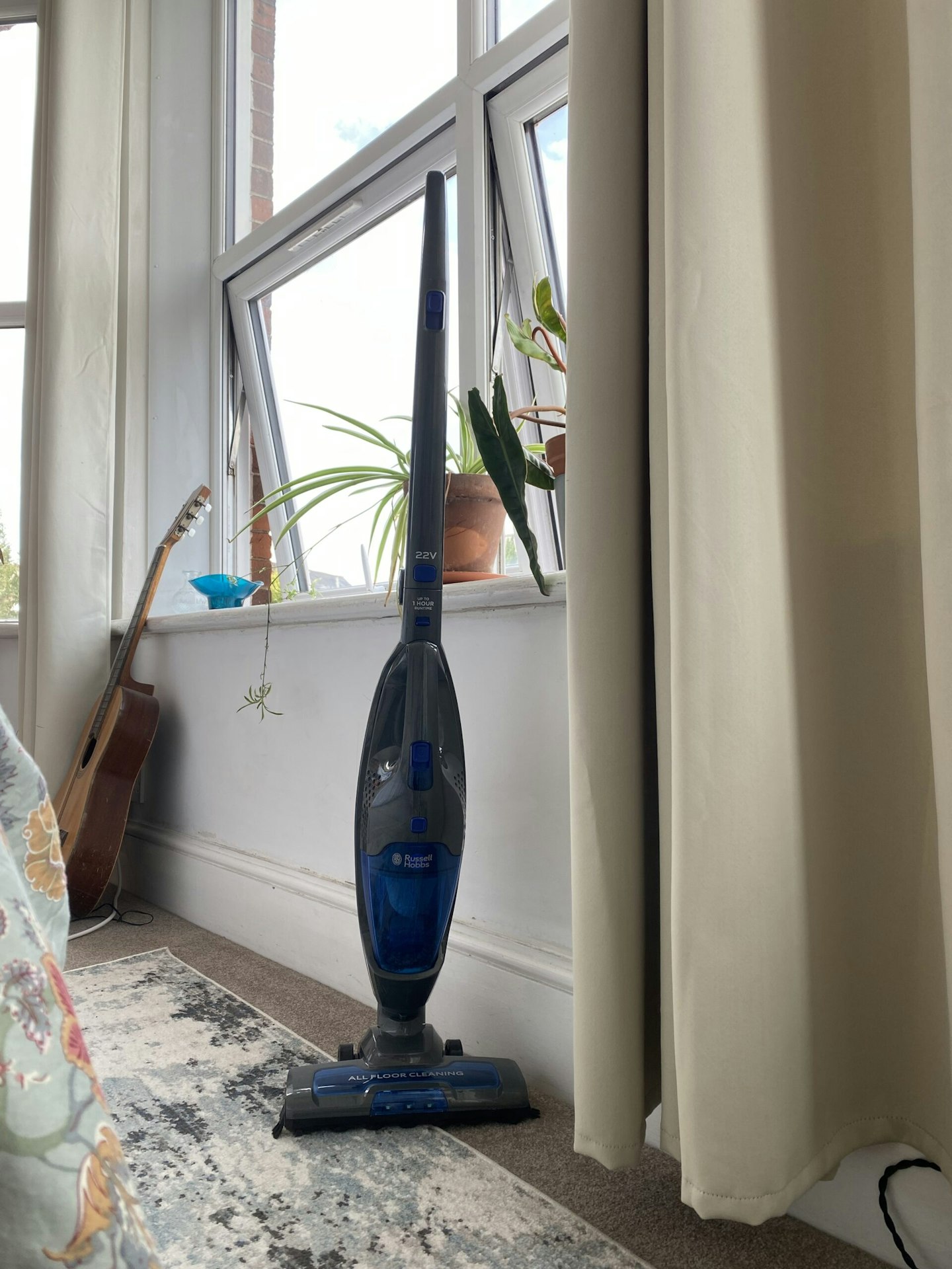 photo of the Russell Hobbs Cordless Upright Stick Vacuum Cleaner in a home setting, guitar, green plants and window in the background