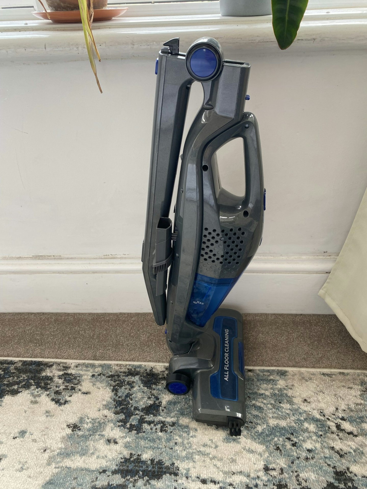 side view of the Russell Hobbs Cordless Upright Stick Vacuum Cleaner with handle collapsed, home setting, skirting board, rug and carpet in the background