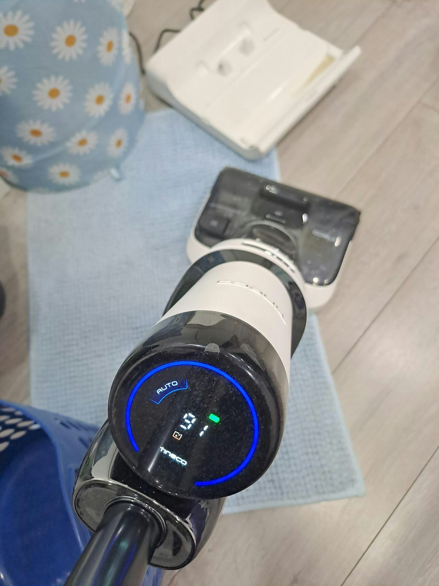 top view of the Tineco Floor ONE STRETCH S6 Cordless Wet Dry Vacuum Cleaner showing the digital display, pale laminate flooring, blue mat, laundry basket and vacuum's charging stand in the background