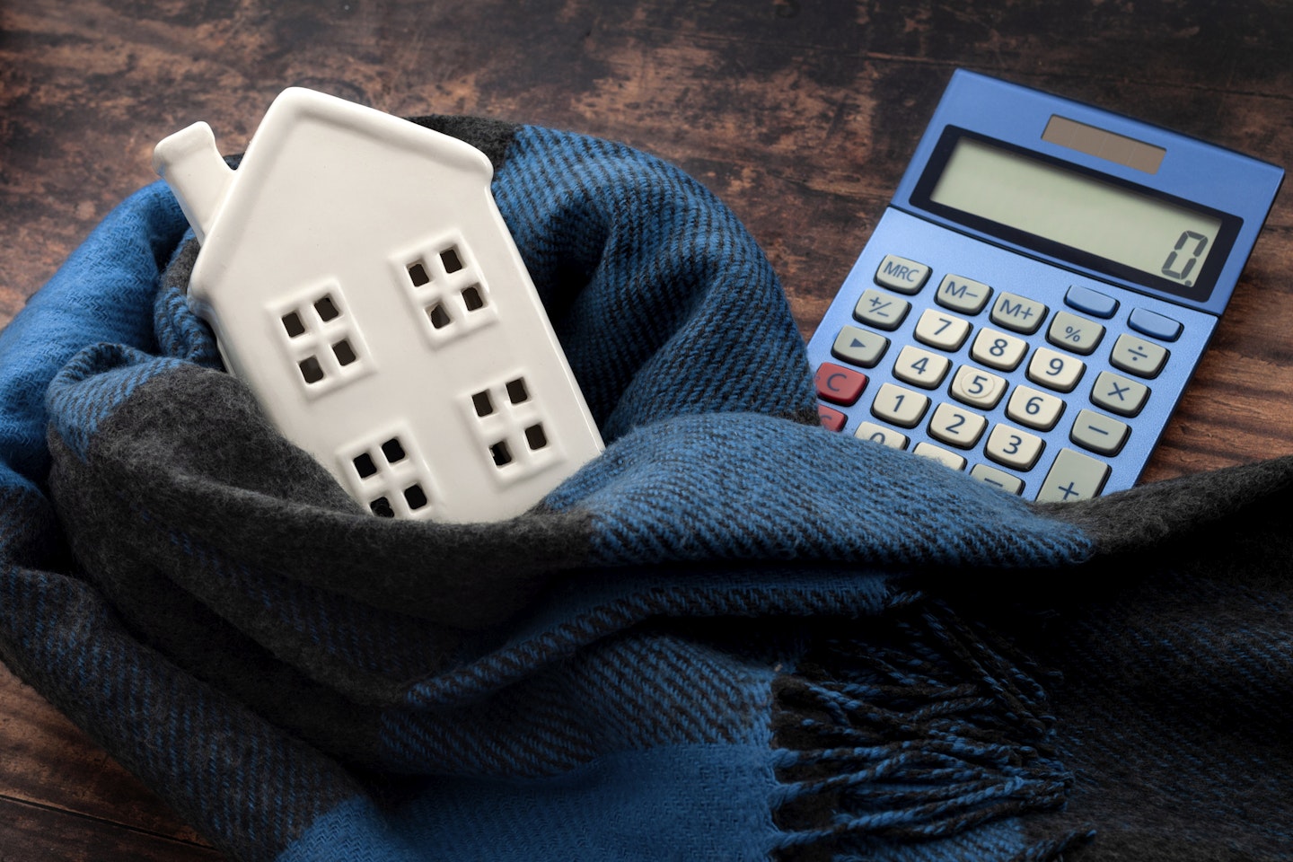 Warming efficiency of a residence and calculate heating costs concept with digital calculator, small house wrapped in warm wool neck scarf on wood background