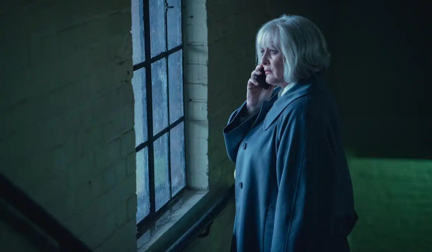 Black Doves Spymaster Reed (Sarah Lancashire) is holding a mobile phone whilst looking into a window. She has a blonde bob and grey coat.