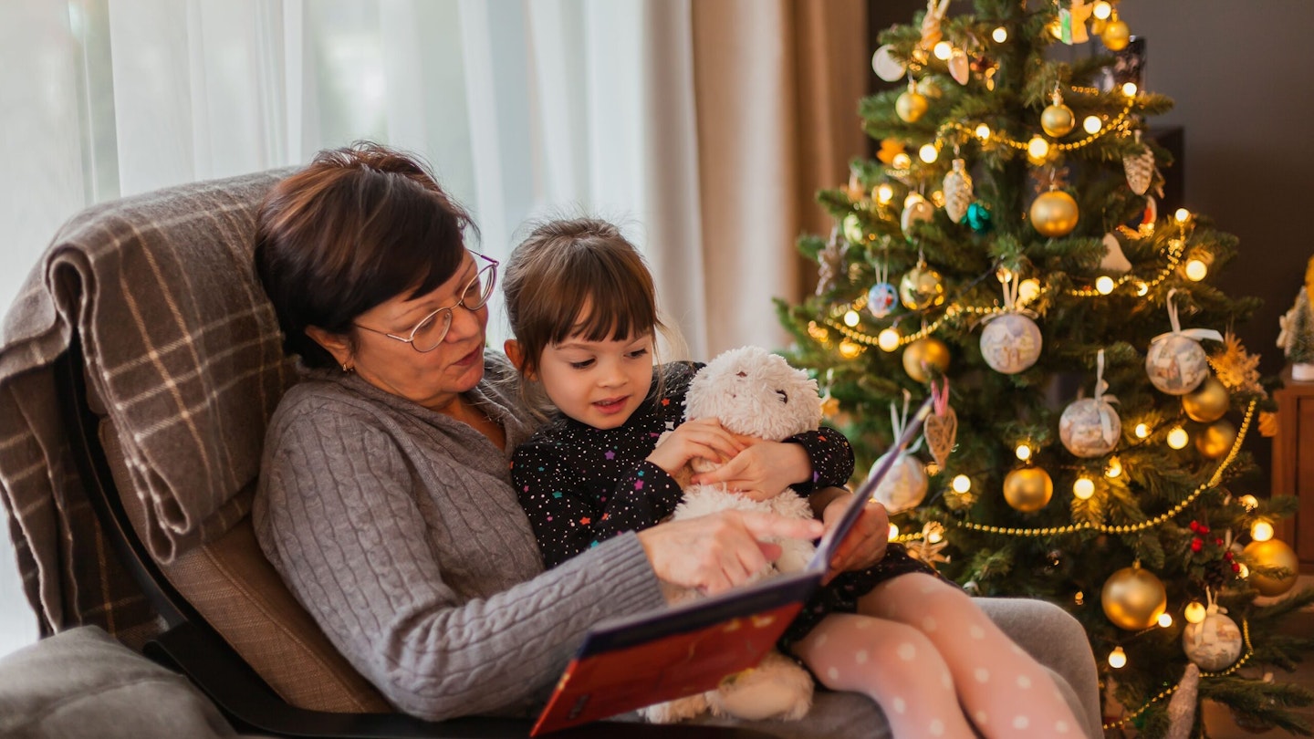 Grandma holding a small girl on her lap as she reads a story to her in front of a Christmas tree