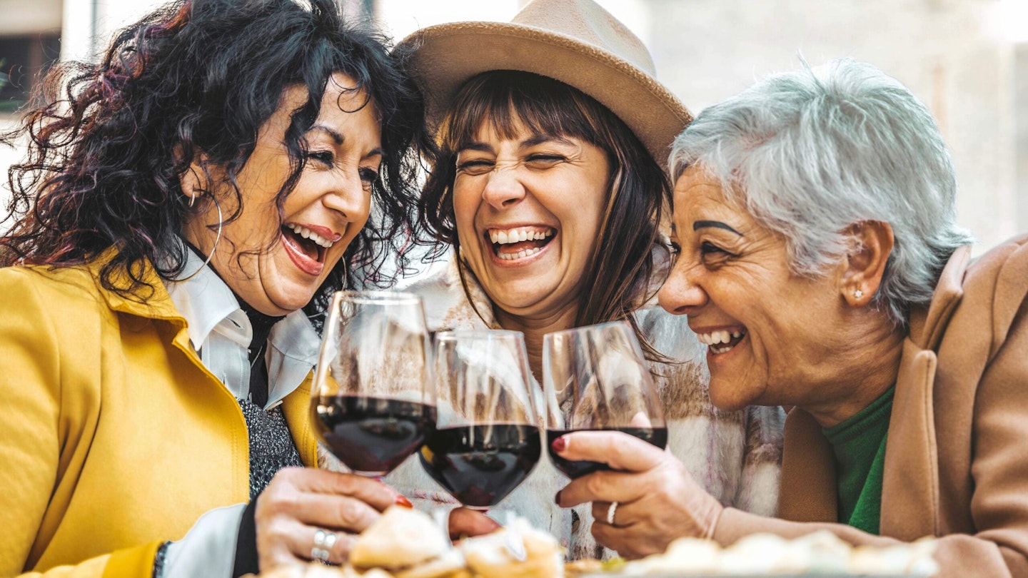 Group of three women with glasses of red wine, laughing.