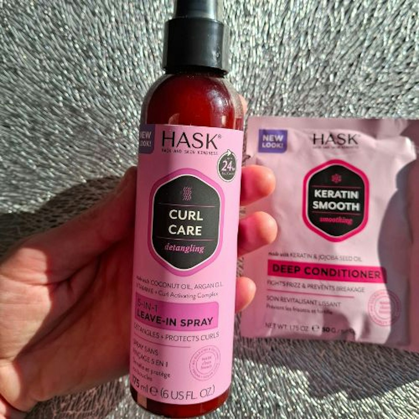 photo of Hask Curl Care Smooth 5-in-1 Leave-in Spray in tester's hand, plus Keratin Smooth Deep Conditioner sachet in the background, on a bright silver grain background