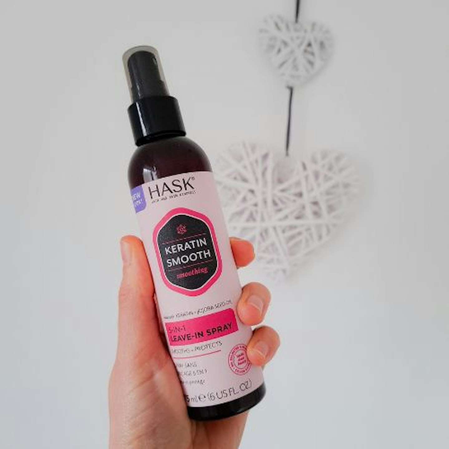 photo of a bottle of Hask Keratin Smooth 5-in-1 Leave-in Spray in tester's hand, white background with a heart wall hanging