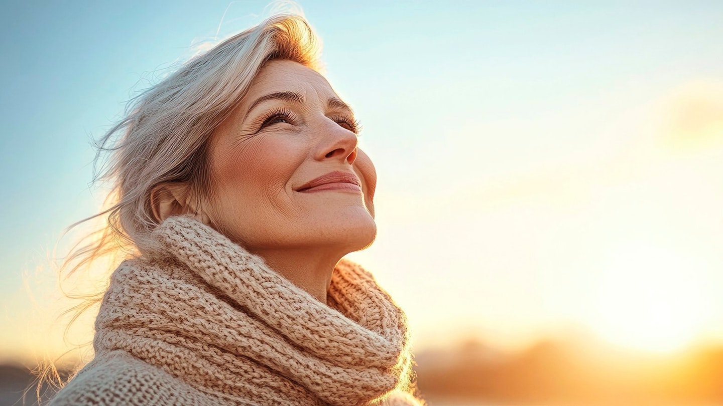 Portrait of a beautiful and happy mature woman in a warm jumper enjoying the winter sun set against a blue sky