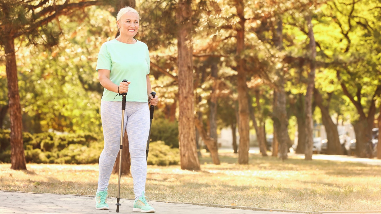 Woman with blonde hair in light green t-shirt and grey leggings, using walking poles