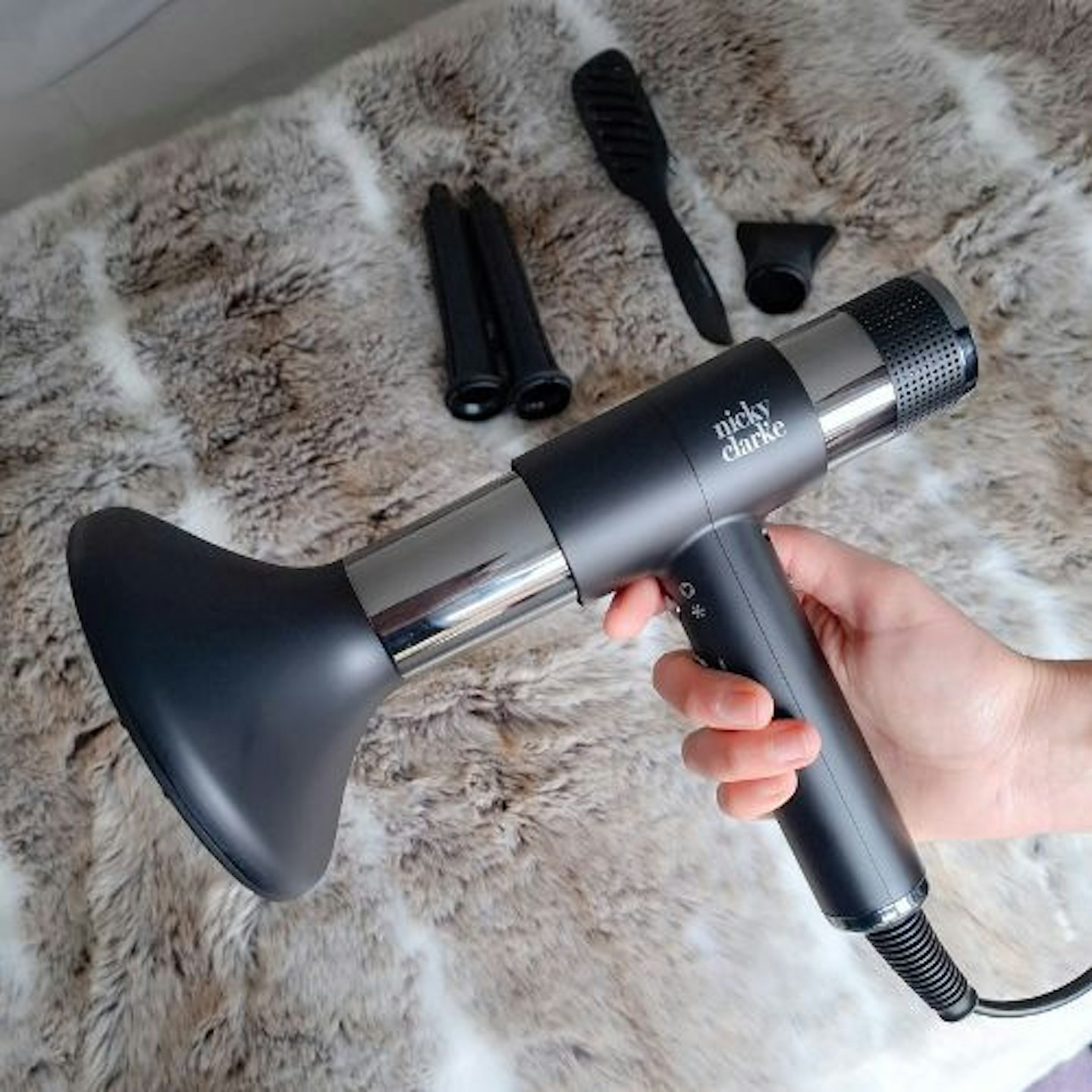 Tester holds the Nicky Clarke AirStyle Pro Infrared & Ionic Hair Dryer & Styler with additional tools in the background on a faux fur throw