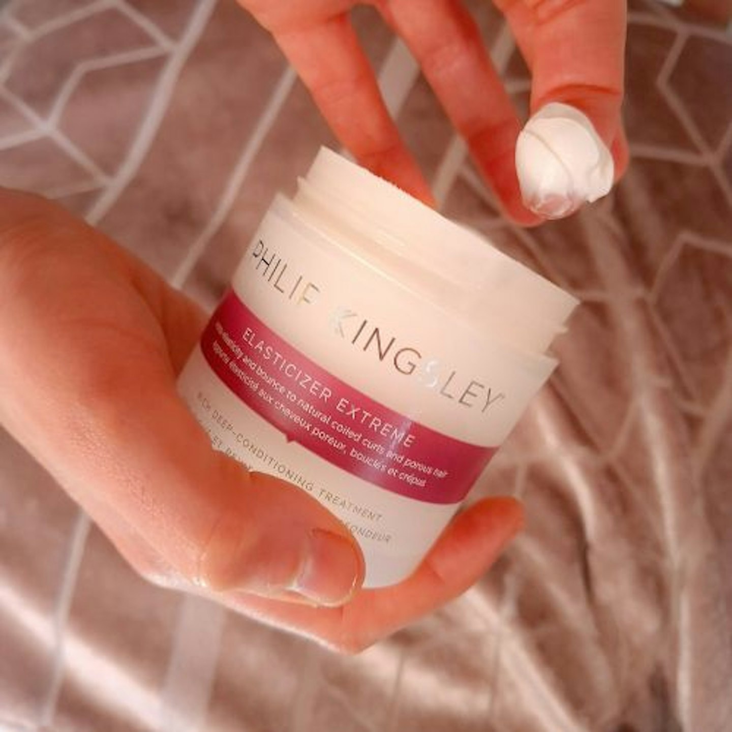 close up of a pot of Philip Kingsley Extreme Elasticizer with some of the cream on the tester's finger, showing it's thick consistency. Pink velour patterned background