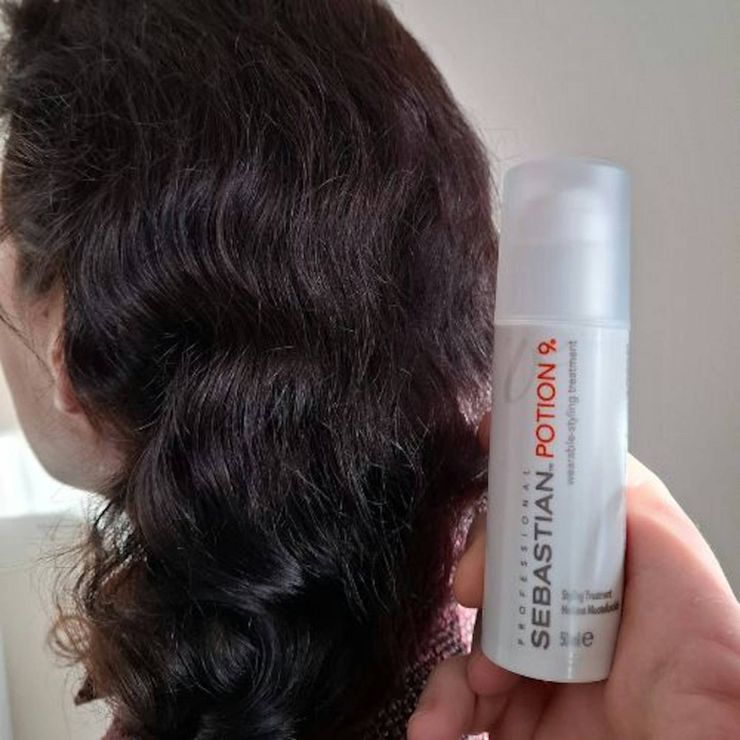 back of tester's dark brown curly hair, showing shine and defined waves, with tube of Sebastian Professional Potion 9 Leave-In Styling Conditioner held next to hair