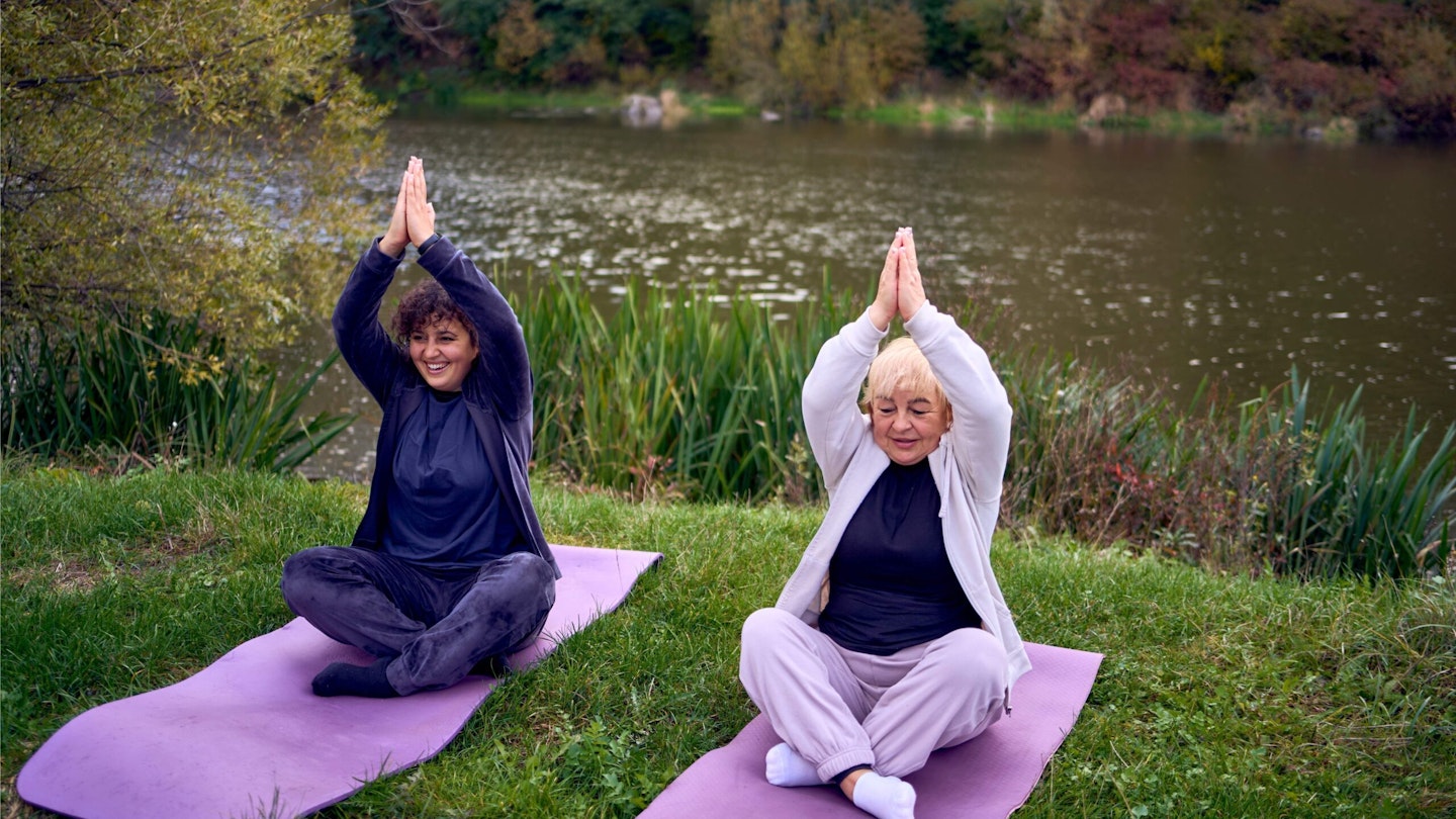 Two women sitting outside on the grass on yoga mats with arms in a prayer position above their heads