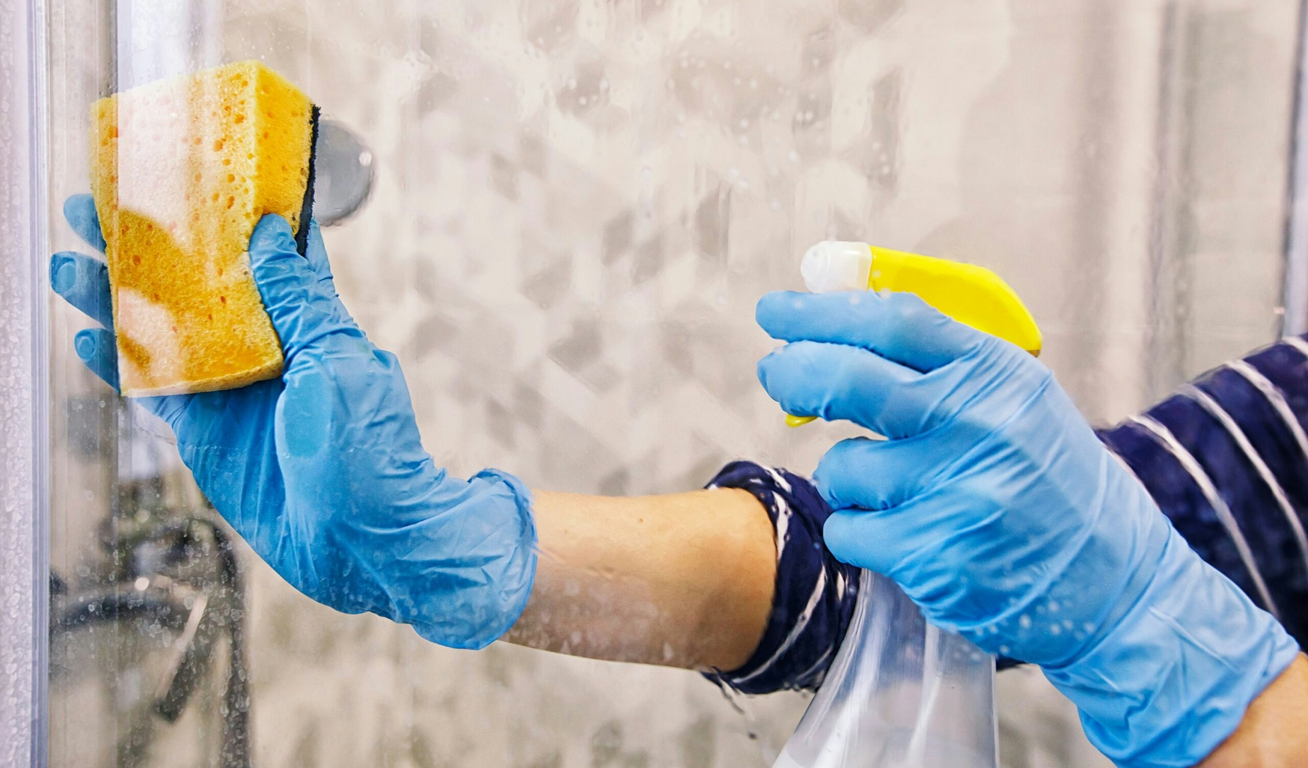 A young woman cleaning the glass in the shower stall using a scrub and spray