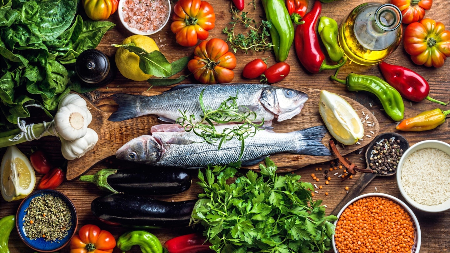 Raw uncooked seabass fish with vegetables, grains, herbs and spices on chopping board over rustic wooden background