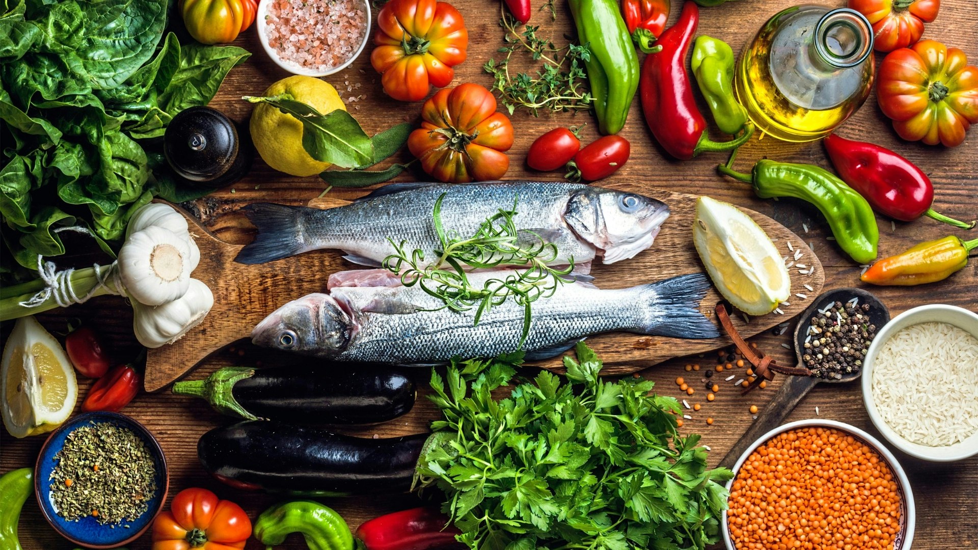 Raw uncooked seabass fish with vegetables, grains, herbs and spices on chopping board over rustic wooden background