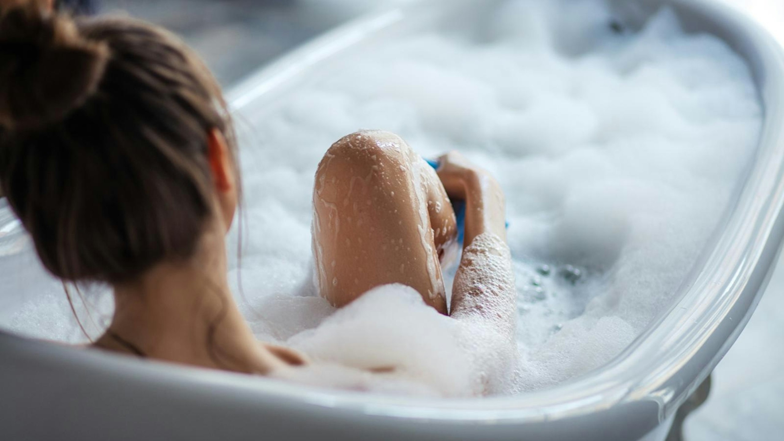 Female massaging her legs with sponge in the tub, back view shot