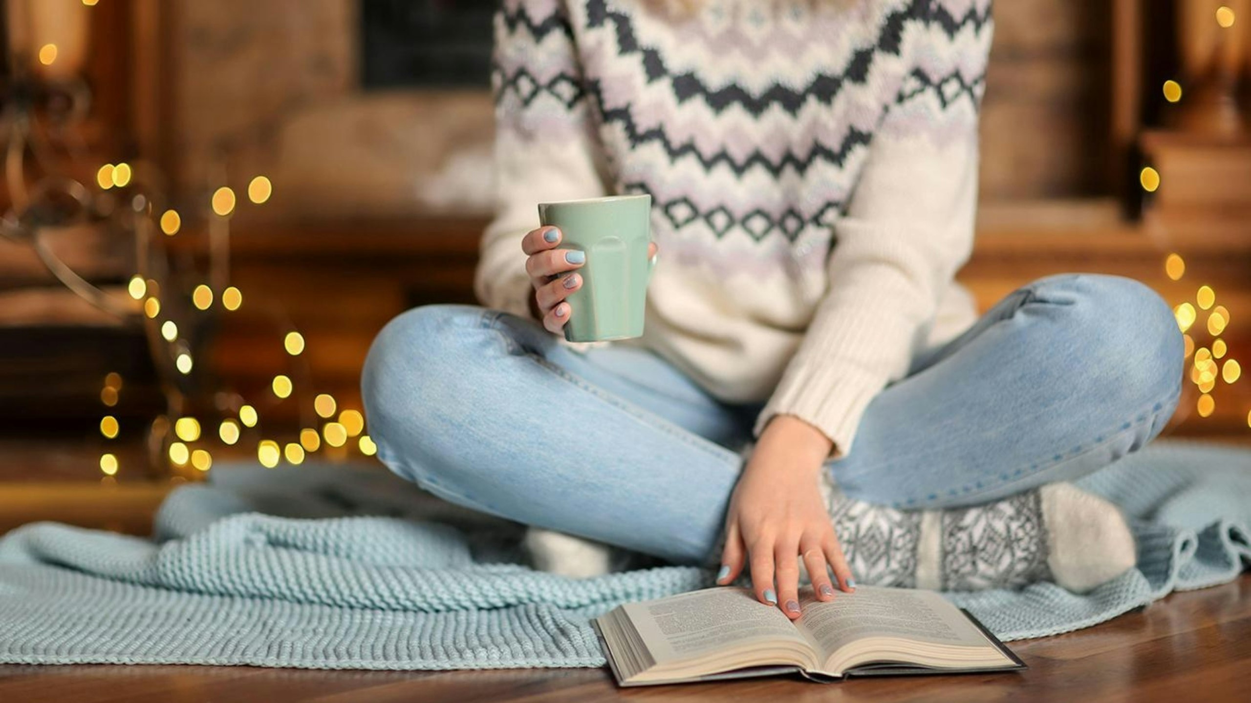 Young woman with cup of tea relaxing while reading book in living room decorated for winter holidays