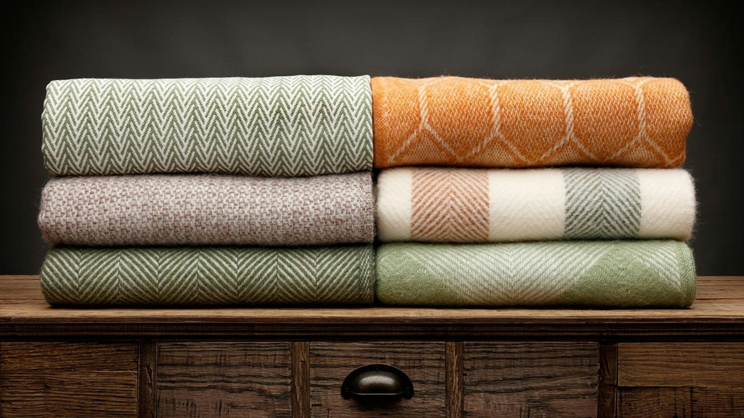 6 luxury throws, folded up and shot on a wooden sideboard, above a draw, with a grey background