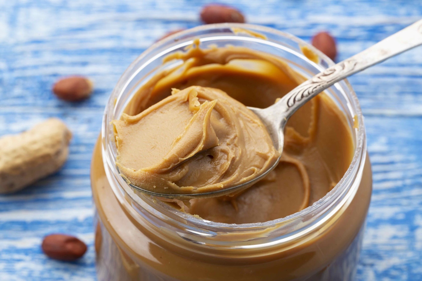 Peanut butter in an open jar and peanuts in the skin are scattered on the blue table