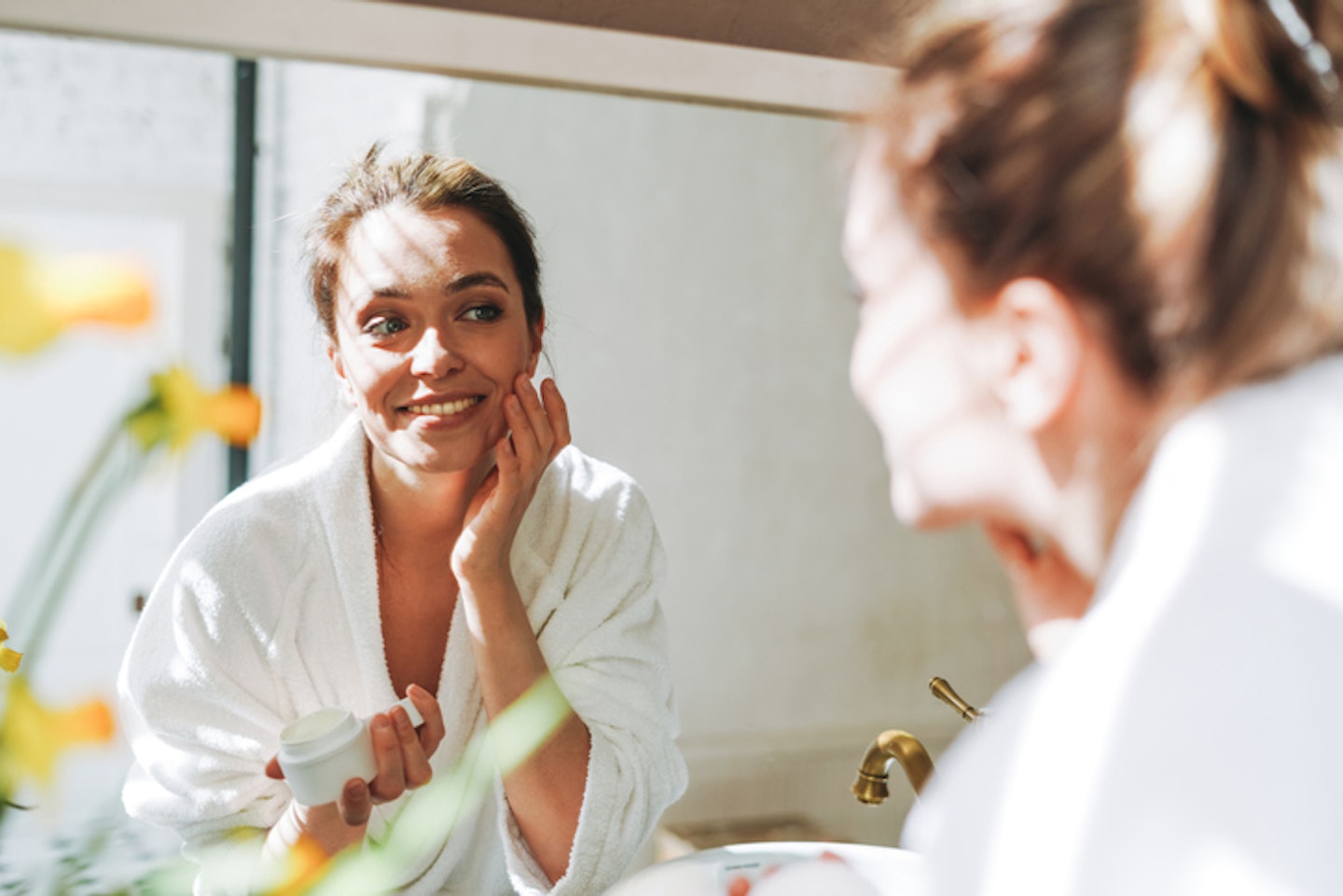 Young Woman In White Bathrobe Put Cream On Clean Fresh Skin Face Near Mirror In Bathroom