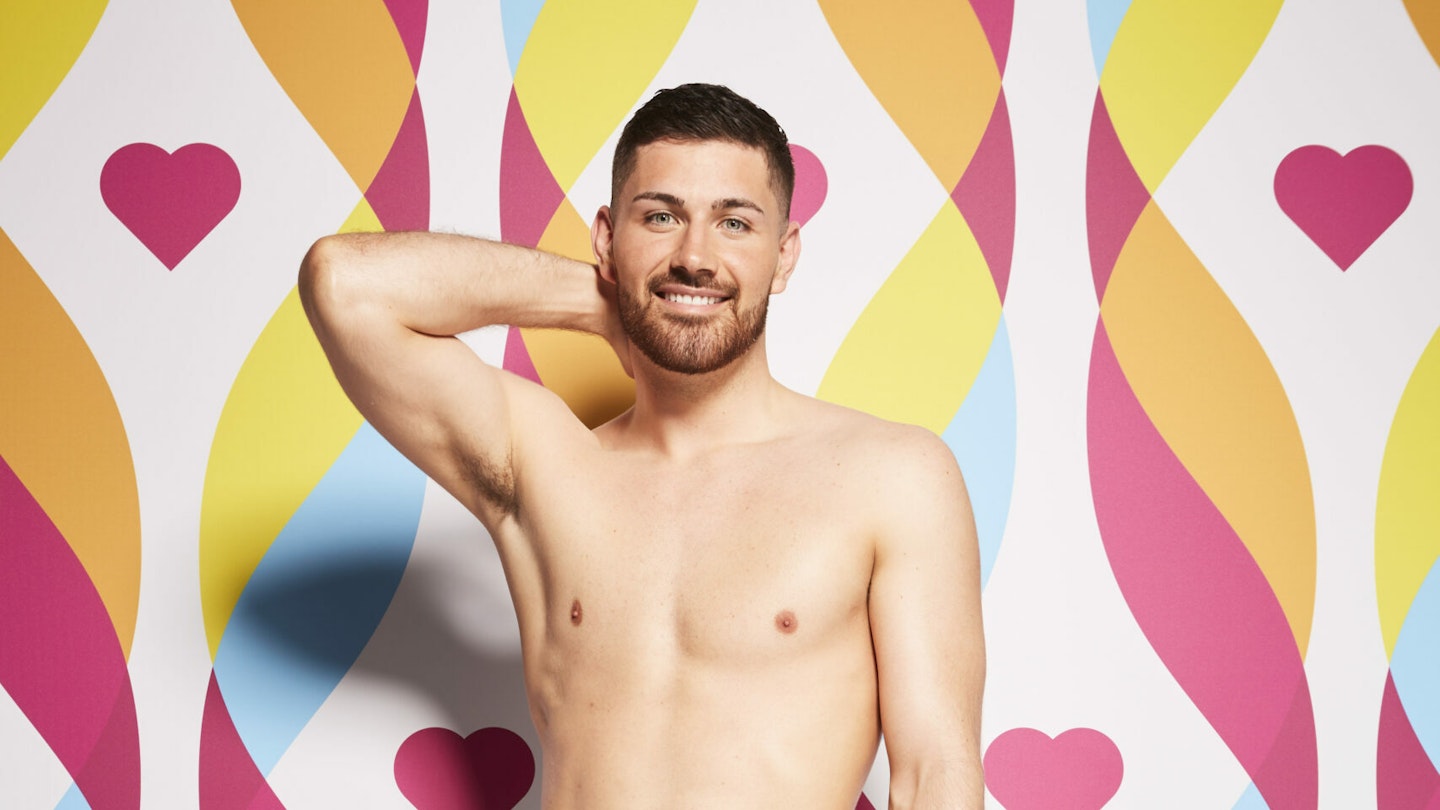 Scott Van Der Sluis topless against a bright patterned background, smiling with his hand being his head.