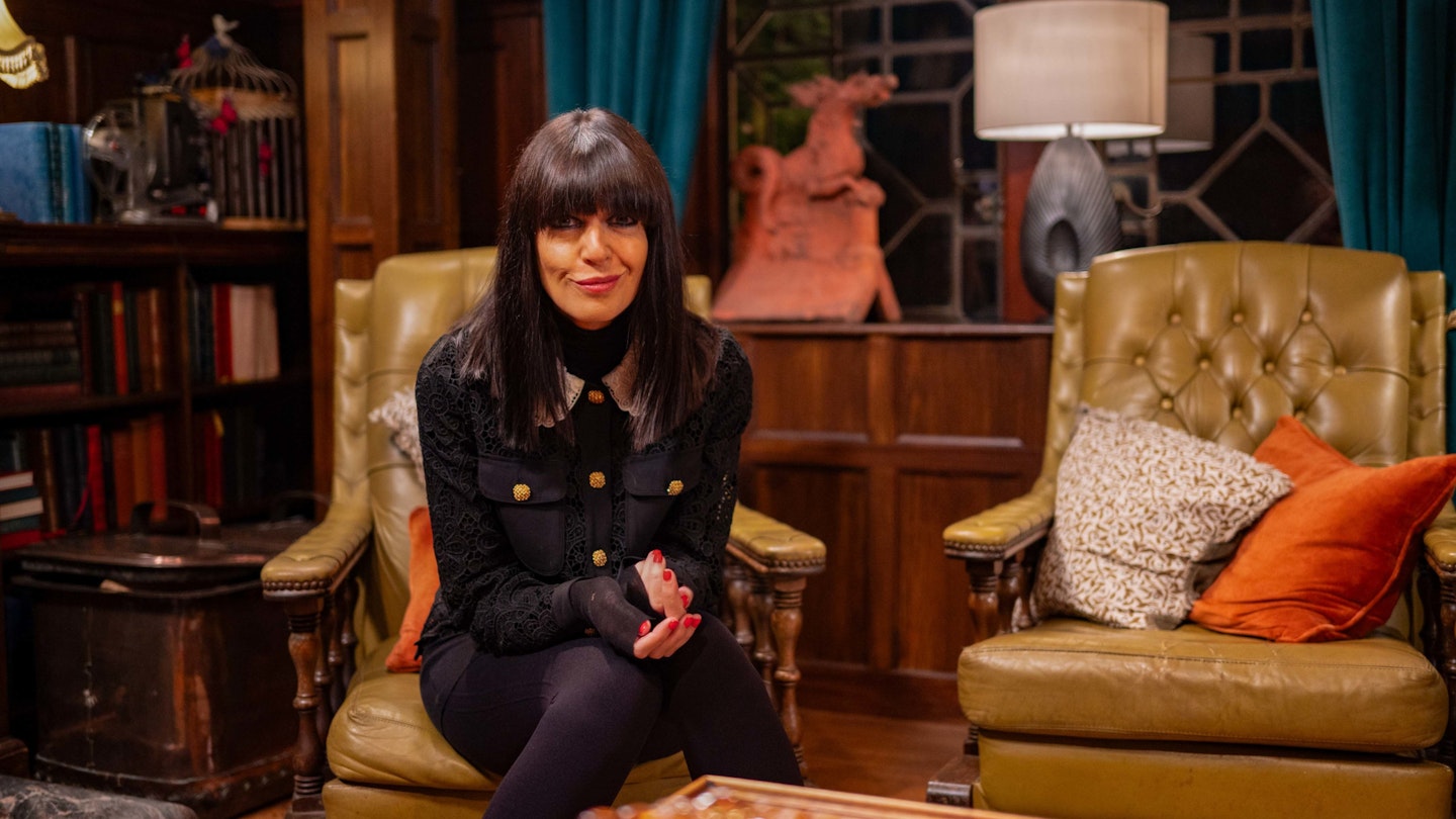 Claudia Winkleman, a slim, white woman with dark hair, sits on a leather armchair with a mischievous grin on her face – on the set of the BBC Show, The Traitors.
