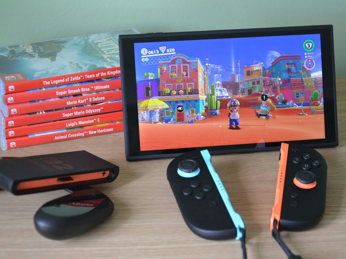 Nintendo Switch 2 in upright position with kickstand, Joy-Con attached in handheld mode – playing Mario Odyssey in Sand Kingdom; original Switch game cases stacked on the left, Joy-Con controller visible bottom left