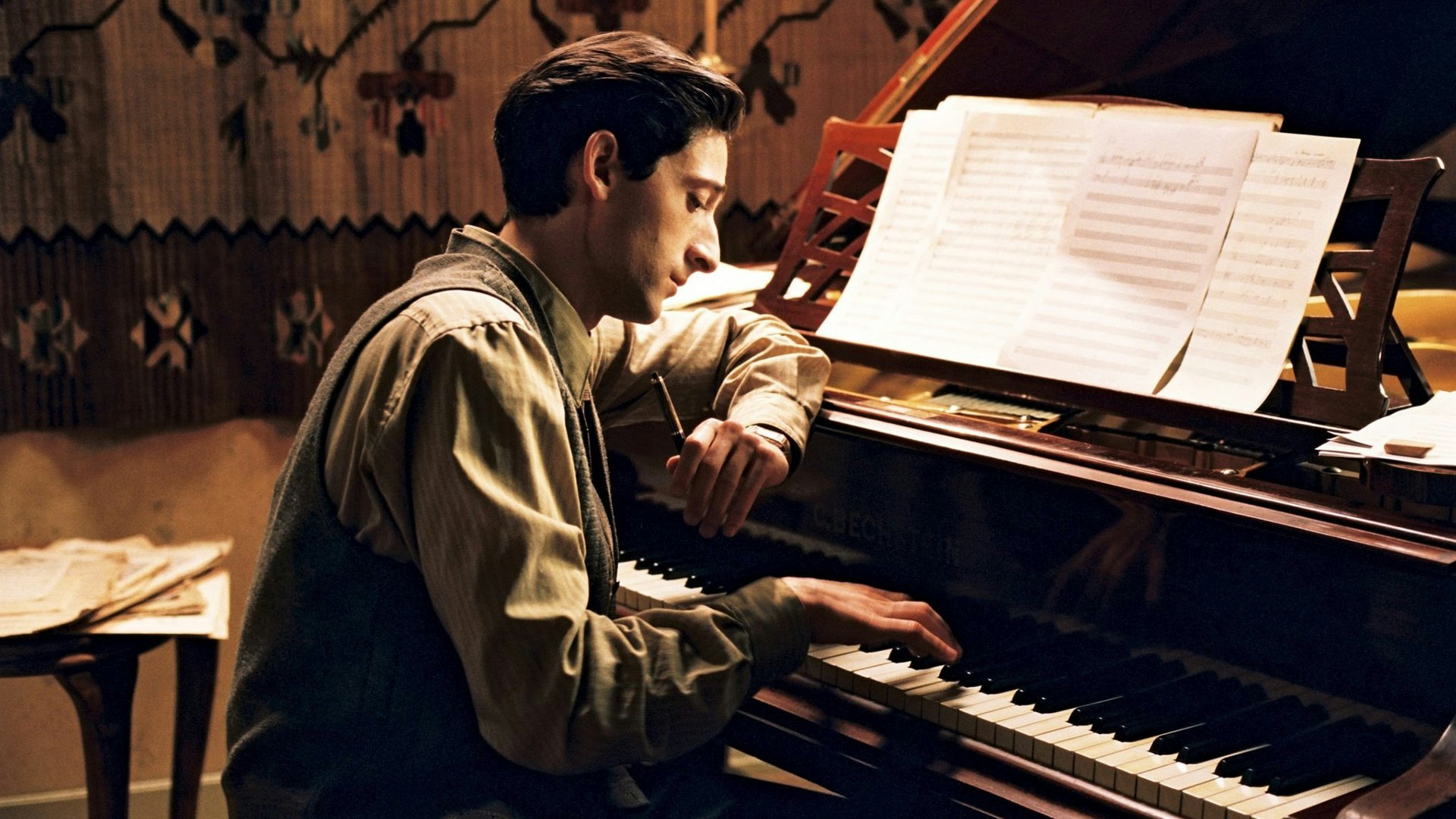 A man with dark hair sits at a piano, looking down at the keys. He is wearing a tan shirt and holding a pen in his left hand, while playing something with his right. There is sheet music on the piano.