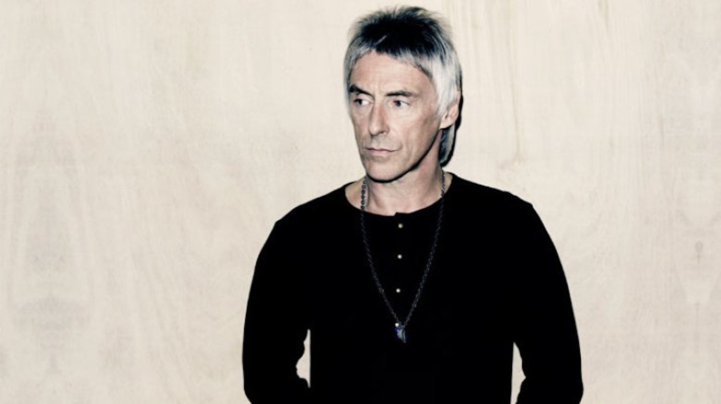 Paul Weller poses in a black top against a white background