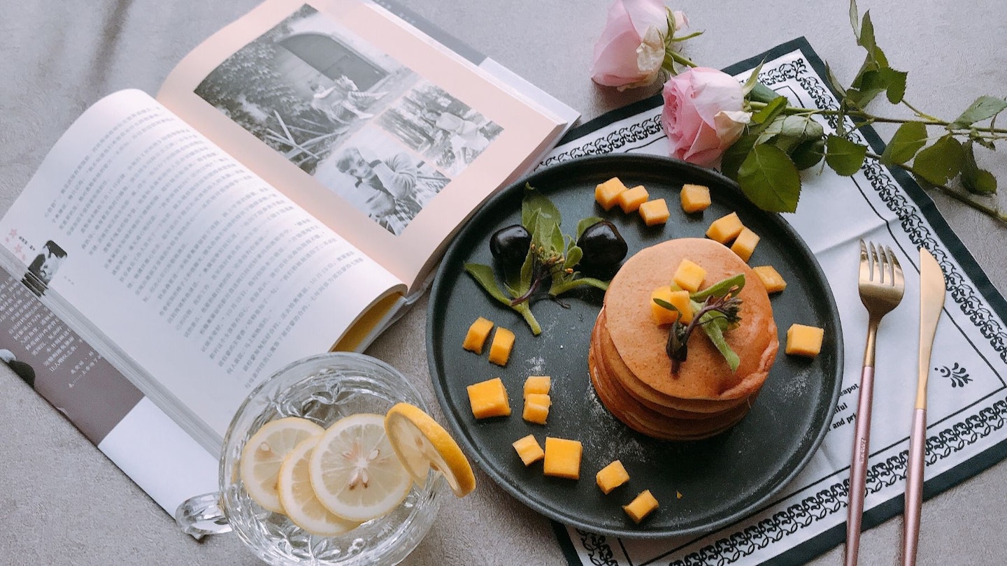 Pancakes with mango on grey background with lemon juice, book, flowers and cutlery 