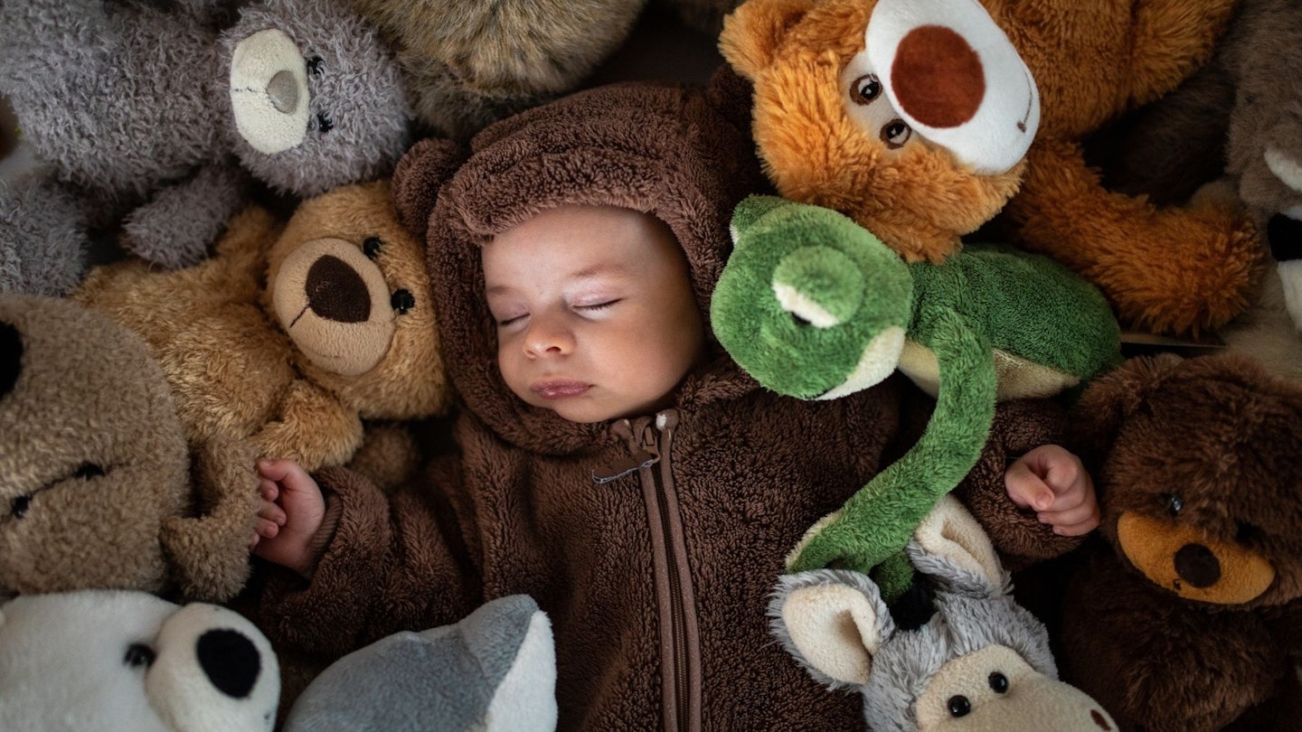 Best teddy bear storage: Directly above of baby boy in bear costume sleeping hidden in stuck of stuffed toys on bed