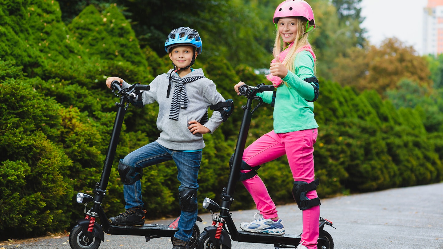 Boy and girl standing on electric scooters wearing protective gear