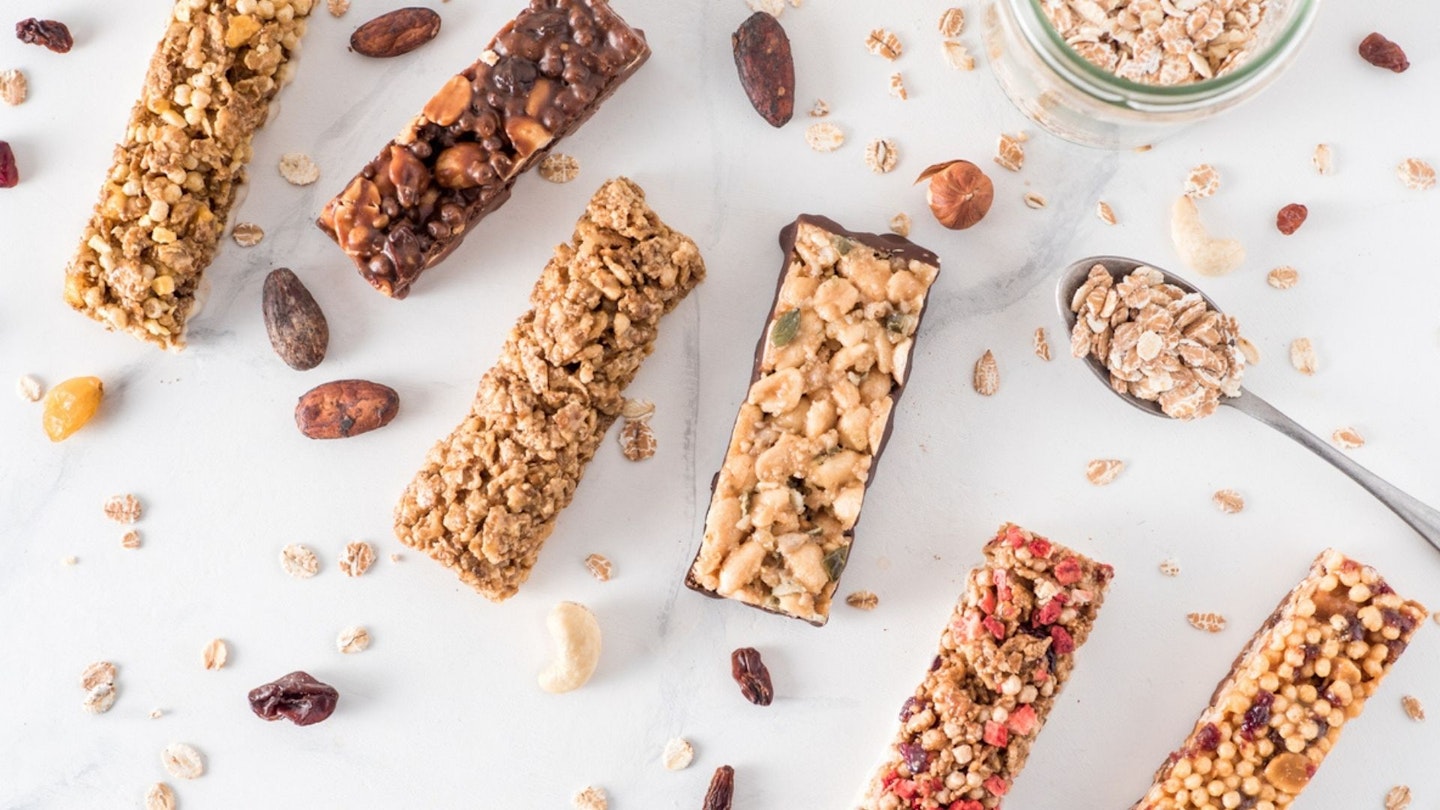 A variety of cereal bars next to a pot of oats and surrounded by fruit and nuts